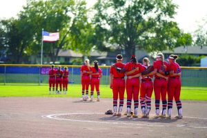 Softball players show team unity during the National Anthem. Players stand in groups based on their field position and the teammates they work most closely with during the game. Grouping 1 is third base, pitcher, catcher, first base. Grouping 2 is short stop and 2nd. Group 3 are the outfielders. Photo by Dawn Meyer